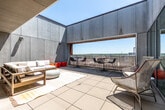 Located at 219 E Grand Ave in Des Moines, Iowa, this rooftop terrace offers a sophisticated outdoor retreat with a distinctly modern architectural character. The space is defined by sleek, large-format concrete panel walls that frame an open-air courtyard, creating a semi-enclosed environment that balances privacy with expansive views of the surrounding Des Moines skyline and cityscape.

The terrace is organized into several distinct functional zones. A generous lounge area anchors one section of the space, furnished with a teak-framed sectional sofa dressed in white cushions, a round ottoman, and woven area rugs that add warmth to the large-tile flooring. Vibrant orange accent seating provides a bold pop of color throughout.

A dedicated bar and dining area features a built-in counter with a bold red paneled backdrop, accompanied by multiple bistro-height tables and sleek wire-frame chairs — ideal for casual dining or social gatherings. Woven lounge chairs are also scattered throughout for additional flexible seating.

Floor-to-ceiling glass sliding doors connect the terrace to an interior space, flooding both areas with abundant natural light. Open sightlines over the perimeter railing deliver sweeping views of the Des Moines urban landscape, making this terrace a compelling venue for events, entertaining, or everyday outdoor relaxation.
