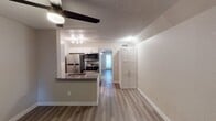 This single-story unit in Phoenix, Arizona offers a straightforward, functional layout with modern finishes throughout. The open-concept living and dining area is finished with wood-look luxury vinyl plank flooring, neutral gray walls, and a textured ceiling. A ceiling fan with integrated lighting serves both the living area and an adjacent space, promoting airflow well-suited to the Arizona climate. Natural light enters through a window fitted with horizontal blinds, and a glass-panel door provides additional brightness to a secondary area of the unit.

The kitchen is positioned along one side of the main living space and features a peninsula with a dark countertop and an integrated sink, offering a casual breakfast bar that opens to the living area. White shaker-style cabinetry lines the walls, complemented by a white subway tile backsplash and stainless steel appliances, including a refrigerator, range with glass-top cooktop, over-the-range microwave, and dishwasher. Track lighting illuminates the workspace effectively.

A hallway leads to the bathroom, which includes a floating vanity with a framed mirror, wall-mounted lighting, and a full-size soaking tub with a tub/shower combination. A built-in storage unit with panel doors is also accessible from the hallway, providing practical organizational space throughout the unit.
