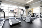 This fitness center is a comprehensively outfitted facility designed to support a wide range of workout routines. The space is finished with neutral-toned walls, crown molding, and rubber flooring throughout, creating a clean and professional athletic environment. Large multi-pane windows along one wall flood the cardio area with natural light and offer views of an outdoor pool and surrounding grounds, making workouts feel open and energizing. Multiple treadmills and an elliptical trainer are positioned to take full advantage of these views, accompanied by a ceiling-mounted flat-screen TV for entertainment during cardio sessions. A dedicated strength training zone features a full multi-functional power rack, a heavy punching bag, and a well-stocked dumbbell rack with a range of weights, alongside an adjustable weight bench for versatile resistance training. A separate area houses spin bikes and additional cardio equipment, while mirrored walls enhance the sense of space and allow for form monitoring. A mini trampoline adds a fun, low-impact training option. The facility is well-lit with flush-mounted overhead lighting, features a first aid station, and is clearly identified with a posted Fitness Center placard. French doors and interior doors provide easy access throughout the space, rounding out a facility built for serious, well-rounded fitness use.