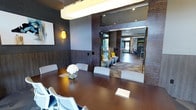 This single-floor space presents a polished, contemporary environment designed for both professional meetings and relaxed gathering. The centerpiece is a spacious conference room featuring a large, rich dark-wood oval table surrounded by modern white chairs, accented by a bold overhead pendant light. Wall sconces with geometric amber shades add warmth against deep gray and wood-paneled walls, while abstract artwork contributes a curated, sophisticated touch. A fresh floral centerpiece and notepads on the table signal a space ready for productive use.

Glass-paneled sliding doors connect the conference room to an adjacent lounge area, which features low-profile seating with gray upholstery and bright yellow accent pillows, creating a vibrant contrast against the neutral palette. A geometric-patterned floor in the transitional corridor adds visual interest and guides movement through the space.

Beyond the lounge, an open common area is visible, bathed in natural light from large exterior windows and glass entry doors, enhancing the airy feel of the overall layout. The space balances functional design with modern aesthetics, making it well-suited for professional collaboration, community gatherings, or casual meetings in an inviting, thoughtfully designed setting.