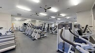 This expansive fitness center offers a thoroughly equipped workout environment across a single, open floor plan. The space is thoughtfully designed to accommodate a wide range of exercise disciplines, from cardio training to strength and functional fitness.

The cardio zone features a long row of treadmills and elliptical trainers, positioned alongside large windows that flood the space with natural light and provide views of the surrounding greenery. Multiple ceiling fans ensure consistent airflow throughout the facility, contributing to a comfortable workout environment.

The strength training area is stocked with a robust selection of Precor and other commercial-grade machines, including cable systems, plate-loaded equipment, a squat rack, and a comprehensive dumbbell rack. Full-length mirrors line the walls, enhancing the sense of openness while allowing users to monitor their form during exercise.

A dedicated functional training zone includes stability balls, a stationary bike, and additional cable machines, offering versatility for varied workout routines. Wall-mounted televisions are positioned throughout the space for added convenience. The durable rubber flooring throughout provides traction and impact absorption. With its bright white walls, ample natural light from arched and multi-pane windows, and a clean, organized layout, this fitness center delivers a professional and motivating atmosphere.