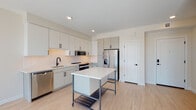 This modern single-floor unit showcases a thoughtfully designed open-concept layout with clean lines and a bright, airy atmosphere. The kitchen is a standout feature, appointed with flat-front cabinetry in a soft grey finish, black hardware accents, and a white subway tile backsplash. Stainless steel appliances — including a French door refrigerator, range, microwave, and dishwasher — complement the contemporary aesthetic, while a mobile kitchen island with a white quartz top and lower storage shelf provides additional workspace and flexibility. Light wood-tone flooring runs throughout the unit, lending warmth and continuity to the space. The open living area is bathed in natural light thanks to expansive floor-to-ceiling sliding glass doors framed in sleek black, which open to a balcony and offer sweeping views of the surrounding cityscape and tree canopy. The bedroom continues the same floor-to-ceiling glazing, flooding the space with sunlight and panoramic outdoor views. The bathroom is finished with large-format white tile, a glass-enclosed walk-in shower, and clean, minimalist fixtures. Recessed lighting throughout and neutral wall tones complete the refined, modern ambiance of this well-appointed space.