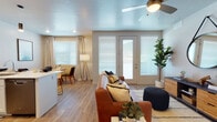 This single-floor residence offers a thoughtfully designed open-concept layout that seamlessly connects the living, dining, and kitchen areas. The living room centers around durable wood-look flooring, a ceiling fan with integrated lighting, a textured ceiling, and a glass-paneled exterior door flanked by windows fitted with horizontal blinds, inviting natural light while offering outdoor views of a hillside landscape.

The kitchen is outfitted with white shaker-style cabinetry, a grey tile backsplash, and stainless steel appliances including a range, microwave, dishwasher, and refrigerator. A peninsula with a white countertop provides bar seating for multiple guests and doubles as a casual dining surface. Sleek pendant lighting hangs above, adding a modern finishing touch.

The bedroom features plush carpeting, a generously sized window with sheer curtains and blinds, and a calm, neutral color palette that creates a restful atmosphere. The bathroom includes a full tub-shower combination with white tile surround, wood-look flooring, and open shelving for storage.

Throughout the space, neutral tones, recessed lighting, and consistent wood-look flooring unify each area into a cohesive, modern living environment.