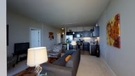 This single-floor space showcases an open-concept design that combines the living and kitchen areas into one cohesive, functional environment. The living area is anchored by a plush gray sofa accented with warm orange throw pillows, complemented by a patterned accent chair that adds visual interest. A large table lamp with a white shade provides warm ambient lighting, contributing to the inviting atmosphere.

The kitchen is well-appointed with dark navy cabinetry contrasting against lighter upper cabinets and white walls, creating a modern two-tone aesthetic. Stainless steel appliances, including a refrigerator, microwave, and range, are seamlessly integrated into the cabinetry. The kitchen also features a stainless steel sink and a small breakfast bar with seating, making it practical for casual dining.

Track lighting runs along the ceiling, supplemented by a pendant light over the kitchen area, ensuring ample illumination throughout. The light-toned flooring unifies the space, while neutral gray walls create a calm, versatile backdrop. A hallway leading to additional rooms is visible, hinting at further functional areas within the unit.