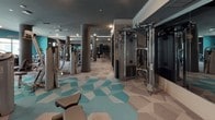 This expansive fitness center is thoughtfully designed to support a comprehensive workout experience across a single open floor. The space is divided into distinct functional zones, seamlessly connected by a striking geometric-patterned carpet in tones of teal, grey, and white that adds a contemporary aesthetic throughout.

The cardio zone is lined with large windows that invite natural light and offer views of the surrounding outdoor area, including a poolside courtyard. This section houses a full row of treadmills, elliptical machines, and rowing machines, providing ample options for cardiovascular training. Structural columns are integrated throughout the space, complementing the modern design rather than interrupting it.

Transitioning deeper into the facility, the strength training area features an extensive collection of professional-grade resistance machines, cable systems, and free weight stations. Floor-to-ceiling mirrors are strategically placed to enhance the sense of space and support proper form during exercise. Recessed and cove lighting, along with ceiling fans, ensure a comfortable environment throughout.

A more enclosed strength and functional training section includes cable crossover machines, benches, and adjustable equipment, accompanied by a wall-mounted flat-screen television. The overall ambiance is sleek and upscale, with a dark and teal color palette, polished finishes, and attentive attention to detail that elevates the fitness experience.
