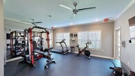 Located at 320 Hannah Todd Pl in Lexington, KY, this fitness facility offers a thoughtfully designed workout environment spread across two exercise rooms on a single floor. The main exercise area is finished with durable dark rubber flooring, neutral gray walls, and crisp white trim, creating a clean and functional atmosphere. Crown molding and recessed lighting add a polished touch, while a ceiling fan ensures comfortable air circulation during workouts. Large windows fitted with white slatted blinds allow natural light to fill the space and offer views of the surrounding exterior. Equipment includes a red and black multi-functional strength training rack with an adjustable bench, a treadmill, a stationary spin bike, a flat weight bench, and a dumbbell rack with a full set of free weights. A large wall-mounted flat-screen TV is positioned for easy viewing during workouts. The secondary exercise room features the same gray and white finishes, recessed lighting, and crown molding, with doors leading to additional utility spaces including what appears to be a restroom. The facility is well-organized, providing users with both cardio and strength training options in a compact yet efficiently utilized space.