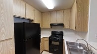 This single-floor space includes a compact yet well-organized kitchen designed for practical, everyday use. The kitchen features light wood cabinetry that lines both the upper and lower walls, providing generous storage throughout. Upper cabinets extend nearly to the ceiling, maximizing vertical storage capacity, while lower cabinets offer additional space beneath the countertops. The countertops are light-colored, complementing the warm tones of the cabinetry. Black appliances anchor the kitchen's aesthetic, including a full-size refrigerator, a gas range with a built-in oven, and a range hood positioned above the stove for ventilation. A stainless steel sink is situated along one wall, adding a functional element to the space. The kitchen is arranged in an efficient L-shaped layout, allowing for ease of movement and workflow. Overhead fluorescent lighting provides bright, even illumination throughout the kitchen. The light-toned flooring ties the space together, creating a cohesive and clean overall appearance.