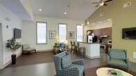 This single-floor space makes a striking first impression with its dramatic vaulted ceilings that soar overhead, creating an airy, expansive atmosphere throughout the open-plan layout. Recessed lighting and a ceiling fan complement the abundant natural light streaming in through multiple tall, framed windows fitted with white blinds, keeping the interior bright and welcoming.

The living area is thoughtfully arranged with comfortable, patterned seating and tasteful wall art, fostering a relaxed yet refined ambiance. Polished concrete-style flooring flows seamlessly throughout, lending a modern, cohesive look to the entire space.

The kitchen is a standout feature, boasting rich dark wood cabinetry, stainless steel appliances including a range and hood, a mosaic tile backsplash, and a peninsula with bar-height seating — perfect for casual dining or entertaining. Pendant lighting above the peninsula adds a warm, inviting glow.

The open connection between the kitchen and living area enhances the sense of spaciousness, making the layout highly functional and social. Overall, the space combines contemporary design elements with a warm, livable feel, offering a well-lit and stylishly appointed environment ideal for comfortable everyday living.