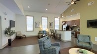 This single-floor space makes a striking first impression with its dramatic vaulted ceilings that soar overhead, creating an airy, expansive atmosphere throughout the open-plan layout. Recessed lighting and a ceiling fan complement the abundant natural light streaming in through multiple tall, framed windows fitted with white blinds, keeping the interior bright and welcoming.

The living area is thoughtfully arranged with comfortable, patterned seating and tasteful wall art, fostering a relaxed yet refined ambiance. Polished concrete-style flooring flows seamlessly throughout, lending a modern, cohesive look to the entire space.

The kitchen is a standout feature, boasting rich dark wood cabinetry, stainless steel appliances including a range and hood, a mosaic tile backsplash, and a peninsula with bar-height seating — perfect for casual dining or entertaining. Pendant lighting above the peninsula adds a warm, inviting glow.

The open connection between the kitchen and living area enhances the sense of spaciousness, making the layout highly functional and social. Overall, the space combines contemporary design elements with a warm, livable feel, offering a well-lit and stylishly appointed environment ideal for comfortable everyday living.