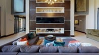 This single-floor amenity clubhouse is designed with a modern, upscale aesthetic that combines comfort and entertainment. The space is anchored by a dramatic lounge area featuring soaring double-height ceilings, a large sectional sofa, and a bold floor-to-ceiling dark wood feature wall fitted with built-in shelving, decorative accents, and a flat-screen television — all illuminated by a striking sputnik chandelier. Light tile flooring flows throughout, contributing to a bright and open feel.

A dedicated billiards area sits adjacent to the lounge, offering a full-sized pool table set beneath pendant lighting, with a built-in upholstered bench and decorative wall shelving providing a refined backdrop. Nearby, a round poker table surrounded by zebra-print chairs occupies its own nook, bathed in natural light from large windows framed by sheer charcoal drapes that overlook lush outdoor greenery and palm trees.

The entrance features dramatic floor-to-ceiling glass doors and windows, flooding the space with natural light and providing direct views of the resort-style pool and surrounding residential buildings. The overall design balances contemporary style with social functionality, making it an ideal gathering and recreation space.