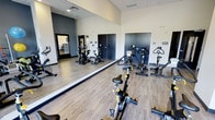 This single-level cycling studio is purpose-built for group fitness, offering a focused and energizing environment for spin workouts. The room is equipped with multiple stationary cycling bikes arranged to allow ample space between participants, ensuring a comfortable and functional workout experience.

A large floor-to-ceiling mirror spans the length of one wall, visually expanding the space and allowing participants to monitor their form during sessions. The wood-look flooring provides a durable yet attractive surface that complements the room's modern aesthetic, while the neutral-toned walls and recessed ceiling panels contribute to a clean, streamlined appearance.

The studio benefits from well-distributed overhead lighting, maintaining a bright and energizing atmosphere suited to high-intensity exercise. Access doors on either side of the mirrored wall provide convenient entry and exit points. The room's layout is practical and uncluttered, keeping the focus firmly on the fitness equipment.

Overall, the space strikes a balance between functionality and contemporary design, making it a well-appointed facility for cycling enthusiasts and group fitness classes alike.