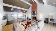 Located at 715 Rice Rd in Ridgeland, Mississippi, this single-story home delivers an impressive open-concept layout anchored by a dramatic great room with soaring vaulted ceilings accented by exposed white beams and a richly textured dark ceiling finish. A floor-to-ceiling brick fireplace serves as the room's focal point, complemented by a wall-mounted television above the white mantel. The generous living area features light tile flooring and is furnished with ample seating, including a large sectional sofa and striped accent chairs, creating a warm and inviting atmosphere. A ceiling fan and pendant lighting enhance both comfort and ambiance throughout the space. The adjacent dining bar area offers counter-height seating with a row of red barstools, seamlessly connecting the living space to the kitchen. The kitchen is well-appointed with white shaker cabinetry, granite countertops, stainless steel appliances including a double oven range and built-in microwave, and rich hardwood flooring. An arched interior doorway leads to additional areas of the home, adding architectural character. Large windows throughout allow generous natural light to fill each room, contributing to the bright and open feel of the entire space.