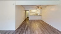 This single-story unit is thoughtfully laid out to maximize functionality across its living spaces. The open-concept living and dining area serves as the heart of the home, featuring wood-look luxury vinyl plank flooring, a ceiling fan with light fixture, and a large window dressed with sheer curtains that allows generous natural light to fill the room. A breakfast bar connects the living area to the kitchen, which is equipped with white cabinetry, ample counter space with granite-style laminate countertops, a white electric range with overhead ventilation, and a sink positioned along the perimeter. Storage is plentiful with upper and lower cabinet configurations throughout.

The bedrooms are carpeted and feature neutral white walls with textured ceilings. Each room is served by windows with vertical blinds that provide views of exterior greenery. The primary bedroom connects directly to a vanity area with a granite-style countertop, white cabinetry, and a large mirror, leading into a full bathroom with a tub-shower combination, toilet, and wood-look flooring. A second bathroom offers similar finishes, including a vanity with under-sink storage. The overall aesthetic is clean and neutral throughout.