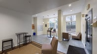 This single-floor lobby serves as a functional and inviting communal space designed to accommodate residents and visitors alike. The layout is open and well-organized, featuring light-toned walls, a drop tile ceiling with recessed and pendant lighting, and durable tile flooring throughout the main area accented by area rugs in the lounge zones.

The space is divided into distinct functional areas. A bright lounge section near the large street-facing windows is furnished with upholstered armchairs and a bistro-height table with bar seating, bathed in generous natural light that offers views of mature trees and the surrounding streetscape. Pendant lights hang above the bistro table, adding a warm, welcoming ambiance.

A secondary seating nook provides a quieter retreat, with two houndstooth-patterned accent chairs arranged on a bordered area rug, complemented by a tall side table and stools. Additional lounge seating is positioned near the building entrance door for easy access.

The mailroom area is clearly integrated into the lobby, featuring a full bank of individual mailboxes mounted on the wall alongside a corkboard for community notices, a wall-mounted intercom panel, and a dedicated counter-height workspace. Safety equipment, including a fire extinguisher and alarm system components, is also conveniently located within the space. Overall, the lobby balances practicality with a comfortable, community-oriented atmosphere.