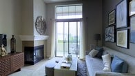 This single-story space centers around a thoughtfully designed living area that balances comfort and elegance. A sleek, modern fireplace with a clean white mantle serves as a focal point along one wall, complemented by a decorative starburst mirror that adds an artistic touch to the neutral, grey-toned walls. Opposite the fireplace, a plush sofa with accent pillows in muted tones anchors the seating arrangement, paired with a tufted ottoman that doubles as a coffee table, styled with decorative accessories.

Floor-to-ceiling sliding glass doors dominate the far wall, flooding the room with abundant natural light and providing direct access to an outdoor balcony. Sheer white curtains frame the doors, softening the transition between indoors and out. The balcony beyond offers greenery and open views, enhancing the airy atmosphere of the interior. A plush area rug grounds the seating area, adding warmth and texture to the light-toned flooring. The overall ambiance is calm and refined, combining contemporary design elements with a welcoming, residential feel.
