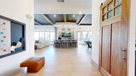 Located at 1600 LPGA Blvd in Daytona Beach, Florida, this single-story community clubhouse offers a thoughtfully designed gathering space that blends modern aesthetics with versatile functionality. The open main area features soaring tray ceilings with dark-painted recessed panels, warm wood beam accents, and statement pendant lighting that creates an inviting, upscale ambiance. Large floor-to-ceiling windows flood the space with abundant natural light and provide views of the surrounding outdoor amenities, including a pool and landscaped grounds.

A centerpiece wet bar with a striking textured tile backsplash, white quartz countertops, and bar-height seating with modern stools serves as the social hub of the space. The adjacent kitchen area features light blue cabinetry, stainless steel appliances, and a large wall-mounted TV, making it well-equipped for events and gatherings. Polished large-format tile flooring runs throughout the main areas, lending a clean, contemporary feel.

The game room section houses both a pool table and a full-length shuffleboard table, offering entertainment options alongside comfortable seating. A sun-drenched dining area with a glass-top table and floor-to-ceiling windows provides an elegant setting for smaller gatherings. Cozy cafe-style seating nooks with marble bistro tables and curated artwork complete the space, ensuring multiple distinct zones for socializing and relaxation.