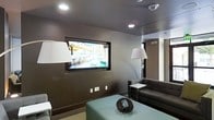 This single-floor space presents a clean, modern aesthetic defined by a neutral palette of deep browns, warm grays, and soft whites. The lounge area serves as the focal point of the space, anchored by a large flat-screen television mounted flush within a recessed wall niche, creating a streamlined, built-in look. Recessed ceiling lighting illuminates the room evenly, complemented by classic white floor lamps that add a warm, ambient glow. Seating is arranged thoughtfully around a light blue-toned coffee table, which introduces a subtle pop of color against the darker surroundings. A dark upholstered sofa and a matching armchair provide comfortable seating, while a compact side table with a white surface offers a functional accent piece. The walls feature a rich, dark brown lower panel paired with lighter gray upper tones, contributing to the space's sophisticated, layered feel. A wall-mounted climate control unit is visible, ensuring comfort throughout. The overall ambiance is polished and contemporary, making the space well-suited for relaxed gatherings or quiet use, with an urban backdrop visible through the television screen hinting at a city setting.