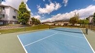 Located in Roy, Utah, this outdoor tennis court is a standout recreational amenity set within a well-maintained residential community. The court features a vibrant blue hard-court surface with crisp white boundary lines and a regulation-height net, offering a professional playing experience in an open-air setting. Surrounded by sturdy chain-link fencing, the court provides a defined and secure play area while maintaining open sightlines to the surrounding grounds.

The setting is lush and inviting, with mature trees lining the perimeter and expansive green lawns extending outward in multiple directions. Multi-story residential buildings are visible in the background, framing the space within an established neighborhood. A covered picnic area with seating is situated nearby, providing a comfortable spot for spectators or those taking a break from play.

Additional community amenities are visible from the court, including a colorful children's playground, further underscoring the family-friendly character of the property. The open sky above allows for abundant natural light throughout the day, making for ideal playing conditions. The combination of a well-kept court surface, scenic surroundings, and proximity to other recreational features makes this tennis court a central and functional gathering space within the community.