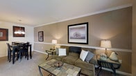 This two-story home offers a comfortable and cohesive living environment defined by classic architectural details and a warm, neutral color palette. The living room serves as a welcoming centerpiece, featuring taupe-toned walls accented by crisp white crown molding and a distinctive chair rail that adds a refined, traditional character to the space. Soft, wall-to-wall carpeting in a complementary beige tone contributes to the room's inviting atmosphere. Ambient lighting from table lamps placed on either side of the seating area creates a warm glow, highlighting the framed artwork displayed on the walls. The layout comfortably accommodates a sectional sofa arrangement paired with a tiled-top coffee table, suggesting a generous and functional floor plan. The clean lines of the ceiling and the well-maintained finishes throughout reflect careful attention to detail. Spanning two floors, the home provides ample separation between living and private spaces, offering both functionality and comfort. The overall ambiance is calm and understated, with design choices that emphasize timeless style and livability throughout.