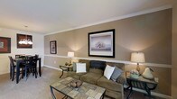 This two-story home offers a comfortable and cohesive living environment defined by classic architectural details and a warm, neutral color palette. The living room serves as a welcoming centerpiece, featuring taupe-toned walls accented by crisp white crown molding and a distinctive chair rail that adds a refined, traditional character to the space. Soft, wall-to-wall carpeting in a complementary beige tone contributes to the room's inviting atmosphere. Ambient lighting from table lamps placed on either side of the seating area creates a warm glow, highlighting the framed artwork displayed on the walls. The layout comfortably accommodates a sectional sofa arrangement paired with a tiled-top coffee table, suggesting a generous and functional floor plan. The clean lines of the ceiling and the well-maintained finishes throughout reflect careful attention to detail. Spanning two floors, the home provides ample separation between living and private spaces, offering both functionality and comfort. The overall ambiance is calm and understated, with design choices that emphasize timeless style and livability throughout.