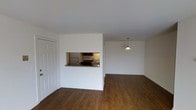 This single-floor space presents a clean, open layout designed for functional everyday living. The main living area is characterized by rich, dark hardwood-style flooring that runs throughout, offering a warm contrast to the crisp white walls and ceiling. A central architectural dividing wall separates the living space from the kitchen while maintaining an open feel through a built-in pass-through window with a granite-style countertop ledge. This design feature allows for easy interaction between the kitchen and living areas, and could serve as a casual breakfast bar.

The kitchen, visible through the pass-through, is equipped with standard appliances including a refrigerator and range, along with dark wood cabinetry and under-cabinet lighting that adds warmth to the cooking space. Adjacent to the kitchen area, a dedicated dining space is defined by a classic pendant light fixture, providing focused illumination for meals. A bi-fold closet door near the entrance offers convenient storage. The overall ambiance is neutral, bright, and move-in ready, with a straightforward layout that maximizes the usable space throughout.