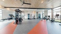 Located at 2701 E Fowler Ave in Tampa, Florida, this expansive fitness center at The Metropolitan offers a thoroughly equipped, single-floor workout facility designed for a wide range of training styles. The space is defined by its striking architectural details, including a warm wood-paneled ceiling with exposed dark steel beams, large ceiling fans, and abundant natural light streaming in through oversized windows along the perimeter walls.

The flooring features a combination of speckled gray rubber and bold orange-accented zones that visually delineate functional training areas from cardio and strength sections. The cardio zone is lined with a row of modern treadmills, ellipticals, and a stair climber, all positioned to take advantage of the natural light and exterior views.

The strength training area includes multiple cable and multi-use machines, dedicated power racks with benches and Olympic weights, and a comprehensive dumbbell rack. A functional training zone showcases a heavy tire, battle ropes, a punching bag, kettlebells, medicine balls, and a plyometric box. Accent walls featuring bold graphic murals add a dynamic visual energy to the space. A refrigerated beverage station and water fountain provide added convenience for users throughout their workouts.
