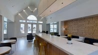 This single-floor space makes an immediate impression with its dramatic vaulted ceilings and bright, open atmosphere. A signature arched window above the main entrance floods the interior with natural light, complementing the recessed lighting and suspended ceiling panels that add a refined, layered look overhead. The centerpiece of the space is a long, sleek reception or service counter finished in crisp white with warm wood base cabinetry, offering both functionality and visual appeal. Yellow metal bar stools provide a pop of color against the neutral palette. A striking textured accent wall clad in warm, dimensional wood panels anchors one end of the room, adding depth and contrast to the otherwise light and airy environment. Seating areas with modern black upholstered chairs are thoughtfully arranged throughout, creating inviting zones for conversation or relaxation. Large windows along the perimeter maintain a strong connection to the outdoors, with views of the surrounding community. The overall ambiance is clean, contemporary, and welcoming, with a careful balance of warm and cool tones, polished surfaces, and carefully chosen greenery that adds a natural, refreshing touch to the interior.