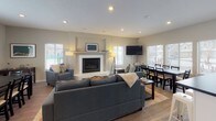 This single-floor community room is thoughtfully designed to serve as a shared gathering space, combining comfort and functionality throughout its open layout. The main living area serves as the focal point of the room, anchored by a tiled fireplace with a clean white surround and a wall-mounted flat-screen television above. Recessed lighting illuminates the space, which is furnished with generous seating including multiple sofas and armchairs arranged around a central coffee table atop a patterned area rug. Large windows fitted with plantation shutters allow ample natural light to fill the room while offering views of the surrounding exterior.

Adjacent to the living area, a dining section provides seating for a group at a dark wood table and chairs, positioned near a window that brings in additional daylight. The kitchen area is fully equipped with stainless steel appliances including a refrigerator, range, and microwave, complemented by white shaker-style cabinetry, light stone-look countertops, and a stainless steel sink. Hardwood-style flooring runs throughout the entire space, tying together each functional zone cohesively. A posted set of clubhouse rules near the entrance indicates this space is a shared amenity available to residents.