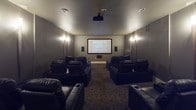 This single-floor private home theater is thoughtfully designed to deliver an immersive cinematic experience. The main screening room is anchored by a large projection screen mounted on the front wall, paired with a ceiling-mounted projector and a full surround sound speaker system with wall-mounted speakers flanking both sides of the room. Recessed lighting and sconce-style wall lights provide soft, dimmable illumination that enhances the theater atmosphere without distracting from the screen.

The seating arrangement features multiple rows of plush black leather recliner chairs, each equipped with built-in cup holders, offering comfortable viewing from every angle. The patterned carpet underfoot adds a classic movie house aesthetic while contributing to the room's acoustic properties.

At the rear of the theater, a well-appointed wet bar area serves as a convenient refreshment station. It includes a round undermount sink, light-colored countertops, white cabinetry, and under-counter appliances including what appears to be a beverage cooler. A framed vintage theatrical poster — a "Don Quixote" Lyceum print — adds a distinguished artistic touch to the space, reinforcing the theater's refined character. The overall ambiance is intimate and upscale, purpose-built for an exceptional private viewing experience.
