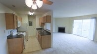 This single-floor residence offers a practical and comfortable layout designed for easy everyday living. The galley-style kitchen is efficiently arranged, featuring wooden cabinetry that provides ample storage, dark countertops with a double-basin sink, a white refrigerator, and a electric range. The tile flooring in the kitchen creates a clean transition into the adjoining living space. A ceiling fan with a light fixture overhead ensures good air circulation throughout the open area. The living room flows naturally from the kitchen, offering a generously sized carpeted space bathed in natural light from a sliding glass door that opens to the exterior. A classic fireplace serves as a focal point in the living area, adding warmth and a welcoming ambiance to the room. The neutral color palette throughout — from the light beige walls to the soft carpeting — creates a calm, move-in-ready atmosphere. A hallway visible from the kitchen suggests additional rooms beyond, contributing to the overall functional design of this well-maintained single-level home.