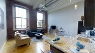 This 1-bedroom, 1-bathroom residence showcases a striking industrial-chic aesthetic rooted in its loft-style architecture. The open-concept living area immediately draws the eye upward to soaring ceilings adorned with white-painted wood planking, exposed structural beams, and a distinctive silver ventilation duct — hallmarks of a thoughtfully preserved industrial conversion. Tall, arched windows along the exposed brick wall flood the space with abundant natural light, highlighting the warm tones of the hardwood flooring throughout.

The living area offers generous room for furnishings, as demonstrated by the seating arrangement anchored by a cowhide area rug, complemented by a dedicated dining nook with acrylic ghost chairs — a modern contrast to the raw brick backdrop. A pendant fan fixture adds both function and style overhead.

Transitioning seamlessly into the kitchen area, a granite-topped island serves as both a prep surface and casual dining counter, providing additional workspace and separation between the kitchen and living zones. The overall ambiance balances rugged, historic character with clean contemporary finishes, creating a distinctive and inviting urban living environment.