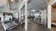 Located at 10 Kialoa Ct in Newport Beach, California, this single-story fitness facility offers a generously sized workout environment organized across multiple functional zones. The space is defined by its distinctive architectural character, featuring vaulted ceilings with exposed white-painted timber trusses in the cardio area, and a coffered ceiling structure with painted steel beams throughout the strength training sections, lending the facility an open, airy feel.

The cardio zone showcases the most architectural detail, with soaring vaulted ceilings, a decorative fireplace, large multi-pane windows on multiple walls, and abundant natural light. Treadmills, elliptical trainers, and a ceiling fan contribute to a comfortable environment for cardiovascular exercise.

The primary strength training area features mirrored walls that amplify the sense of space, dumbbell racks, adjustable benches, cable machines, and a multi-station cable system. Rubber flooring throughout provides durability and safety. A secondary weight room with wood-paneled walls houses free weight benches, a cable tower, and medicine ball storage.

French doors and multiple windows connect the interior to the surrounding Newport Beach environment, allowing natural light to filter through. The facility's layout and architectural details create a practical yet characterful training environment.