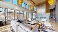 This thoughtfully designed amenity space spans a generous single floor, offering a seamless blend of contemporary architecture and upscale finishes throughout. The centerpiece is a dramatic double-height living and lounge area bathed in natural light from expansive floor-to-ceiling windows, providing sweeping views of the surrounding neighborhood. Striking amber chandelier clusters cascade from the soaring ceiling, adding warmth and a bold design statement to the open layout.

A sleek, linear fireplace anchored by a textured stone accent wall serves as a sophisticated focal point, flanked by curated seating areas with plush sectional sofas and artful area rugs. The kitchen and bar area features a long white quartz countertop, wood-toned cabinetry, herringbone hardwood flooring, and a stunning veined stone backsplash, all illuminated by a cluster of globe pendant lights overhead.

The space also includes a private conference room enclosed in glass with dark-framed sliding doors and city-facing windows, as well as a smaller meeting room adorned with a hand-painted gradient accent wall and an artistic floral ceiling fixture. A hallway connects each zone fluidly, ensuring the entire space functions cohesively for both collaboration and relaxed social gathering.