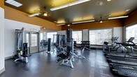 This single-floor fitness center offers a comprehensive workout environment designed to accommodate a range of exercise needs. The space is defined by its bold amber and golden-toned upper walls and ceiling, contrasting with white lower walls, creating an energetic and motivating atmosphere. The polished concrete flooring in a neutral gray tone provides a durable, easy-to-maintain surface throughout.

The main workout area is generously proportioned, housing a variety of strength training machines, including a multi-function cable and weight system, adjustable benches, and a well-stocked dumbbell rack arranged along the windowed wall. Large windows along one side of the room flood the space with natural light, enhancing the open feel of the interior. Ceiling-mounted fluorescent and pendant light fixtures provide additional illumination throughout.

A dedicated accessories area near the entrance features a vertical rack stocked with medicine balls and exercise mats in assorted colors, supporting functional and flexibility training. A wall-mounted mirror expands the visual depth of the main floor. An adjacent room, visible through an interior doorway, contains a mounted monitor, suggesting a supplementary space for stretching or additional activities. The fitness center also benefits from ceiling fans for ventilation and comfort.