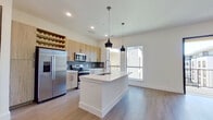 This well-appointed single-floor unit offers a thoughtfully designed layout that maximizes functionality across one bedroom, one bathroom, a kitchen, and a dedicated closet. The open-concept kitchen serves as the centerpiece of the living area, showcasing light wood cabinetry with matte black hardware, a striking herringbone-patterned tile backsplash, and a full suite of stainless steel appliances including a French door refrigerator, electric range, and over-the-range microwave. A sleek white kitchen island with an integrated sink anchors the space and is illuminated by two modern pendant lights, while a built-in wine rack adds a distinctive design touch. Light hardwood-style flooring flows throughout the main living areas, complementing the crisp white walls and bold dark interior doors that lend the unit a contemporary character. The bedroom features soft carpeting, large windows that invite abundant natural light and outdoor views, and a ceiling fan. An adjacent walk-in closet offers multi-tiered shelving and hanging rods for ample storage. The bathroom is finished with wood-toned cabinetry, a white quartz countertop, large-format tile flooring, and a tub-shower combination, all tied together with a clean, modern aesthetic.