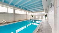 Located on Rue Stanley in the heart of Montréal, this indoor pool facility offers a generously sized, single-level aquatic space designed for both recreation and relaxation. The centerpiece is a large, clearly marked lap pool with vivid turquoise water, framed by smooth tiled pool decking that runs along its perimeter. The architecture is defined by a striking exposed beam ceiling painted in soft blue tones, adding a clean, structured visual character to the space. Walls clad in white ceramic tile run the full height of the room, contributing to the bright, hygienic aesthetic typical of a well-maintained aquatic facility. A row of large windows lines one side of the pool, allowing abundant natural light to flood the interior while offering elevated views of the Montréal cityscape beyond. A dedicated poolside lounge area features a round glass table and chairs, providing a comfortable spot for relaxation. Safety equipment, including a life ring, is visibly mounted along the pool edge. An adjacent entry point provides access to surrounding amenities. Lush tropical potted plants add a touch of greenery, softening the clean, utilitarian lines of the space. Overall, the facility presents a welcoming, well-appointed environment for aquatic activities in the heart of downtown Montréal.