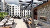 This rooftop amenity deck offers a well-appointed outdoor retreat designed for both relaxation and social gatherings. The space is anchored by a sleek, modern pergola constructed from dark powder-coated steel, providing partial shade over a generously sized lounge area. Beneath the pergola, a curated arrangement of woven all-weather furniture — including sofas, armchairs, and accent tables crafted from warm teak wood — creates an inviting setting for conversation and entertaining. To one side, a built-in outdoor entertainment wall is finished with rich wood paneling and natural stone cladding, housing a large-screen television for open-air viewing. Beyond the covered lounge, rows of cushioned chaise loungers are positioned to take full advantage of open sky and natural sunlight, making them ideal for sunbathing. The deck is finished with light-toned pavers that complement the overall neutral and contemporary color palette. Railing barriers frame the perimeter, offering unobstructed views of the surrounding multi-story residential buildings and distant palm trees, reinforcing the urban yet resort-like character of the space. The overall ambiance is that of a premium, thoughtfully designed outdoor living area that balances comfort, style, and functionality.