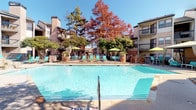The Main Pool area is a well-appointed outdoor amenity space designed for relaxation and recreation. At its center sits a generously sized swimming pool with clear, blue water, complemented by a surrounding concrete and stone deck that provides ample room for lounging. Teal-cushioned chaise lounges and umbrella-shaded seating are arranged throughout the deck, offering both sun and shade options for residents.

Adjacent to the main pool is a distinctive multi-tiered stone water feature, constructed from natural stacked stone with cascading water elements and an integrated spa. This area adds a resort-like ambiance to the space and serves as a visual focal point.

Shaded cabana structures are positioned along the perimeter, each equipped with teal lounge furniture, high-top seating, and retractable privacy curtains, offering a more intimate outdoor retreat. Potted plants and mature trees with seasonal foliage provide natural landscaping throughout.

The pool area is enclosed by an iron fence and is flanked by a brick and stone clubhouse building, which features large windows and direct access to the pool deck. A Texas state flag flies prominently above, anchoring the space with a sense of regional character. The entire area benefits from abundant natural sunlight and open sky views.