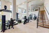 Located at 2700 N Lake Forest Dr in McKinney, TX, this two-story fitness center offers a comprehensive workout facility designed for a wide range of exercise disciplines. The ground floor makes an immediate impression with its soaring double-height ceilings, floor-to-ceiling windows, and an abundance of natural light that floods the open workout areas. Elegant herringbone-patterned hardwood-style flooring anchors the strength training zone, which is equipped with a full dumbbell rack, adjustable benches, cable machines, a squat rack, and a functional training rig. Green turf areas designated for functional and dynamic training are complemented by medicine balls, kettlebells, resistance tools, and a heavy bag. Large mirrors line the walls, enhancing the sense of space. A sleek open-tread staircase with glass railings leads to the second floor, which overlooks the main floor via a modern glass balustrade. The upper level features additional cardio and strength equipment with exterior-facing windows providing views and daylight. Dedicated cardio rooms on both levels house treadmills, ellipticals, and stationary bikes on plush multi-toned carpet tile flooring. Built-in wooden shelving and storage units provide practical organization throughout. Ceiling fans and recessed lighting ensure a comfortable environment across all exercise areas.