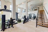 Located at 2700 N Lake Forest Dr in McKinney, TX, this two-story fitness center offers a comprehensive workout facility designed for a wide range of exercise disciplines. The ground floor makes an immediate impression with its soaring double-height ceilings, floor-to-ceiling windows, and an abundance of natural light that floods the open workout areas. Elegant herringbone-patterned hardwood-style flooring anchors the strength training zone, which is equipped with a full dumbbell rack, adjustable benches, cable machines, a squat rack, and a functional training rig. Green turf areas designated for functional and dynamic training are complemented by medicine balls, kettlebells, resistance tools, and a heavy bag. Large mirrors line the walls, enhancing the sense of space. A sleek open-tread staircase with glass railings leads to the second floor, which overlooks the main floor via a modern glass balustrade. The upper level features additional cardio and strength equipment with exterior-facing windows providing views and daylight. Dedicated cardio rooms on both levels house treadmills, ellipticals, and stationary bikes on plush multi-toned carpet tile flooring. Built-in wooden shelving and storage units provide practical organization throughout. Ceiling fans and recessed lighting ensure a comfortable environment across all exercise areas.