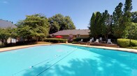 This single-story property is defined by its inviting outdoor amenities and tranquil natural setting. The centerpiece of the exterior is a generously sized swimming pool with crystal-clear turquoise water, divided by lane lines and accessible via built-in entry steps. The pool deck is furnished with multiple lounge chairs, providing ample space for relaxation and recreation under open skies. A distinctive red umbrella adds a pop of color near the building's entrance, offering a shaded spot for outdoor seating. The surrounding landscape is lush and mature, with large deciduous trees creating a verdant canopy that frames the property and provides natural shade and privacy. The building itself is a low-profile, single-story structure with a pitched, dark-shingled roof and earth-toned exterior, blending seamlessly with the wooded surroundings. Well-maintained shrubbery and hedging line the base of the building, contributing to the manicured appearance of the grounds. The overall ambiance is peaceful and resort-like, with the combination of sparkling water, generous greenery, and comfortable outdoor furnishings creating an ideal environment for leisure and outdoor living.