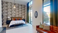 This single-floor space is defined by its dramatic soaring ceilings and an abundance of natural light streaming through expansive floor-to-ceiling windows, offering outdoor views of surrounding greenery. The main room functions as both a sleeping and living area, unified by light-toned tile flooring throughout. A striking botanical-patterned wallpaper serves as a bold focal point behind the bed, complemented by a navy upholstered headboard, coordinating nightstands, and warm orange accent pillows. A ceiling fan overhead adds practical comfort to the generous vertical space. A built-in shelving and storage unit with a mini-refrigerator provides added convenience alongside open wardrobe hanging space. The sitting area features a tufted cognac leather sectional sofa, a cowhide rug, a marble-topped coffee table, and a series of framed botanical artwork arranged on the crisp white walls. Dark green floor-to-ceiling drapes frame the windows elegantly. A light-filled hallway lined with tile flooring connects the main room to a private bathroom, which includes a dark-cabinetry vanity with an undermount sink, dark countertops, and a separate shower, delivering a clean and functional aesthetic.