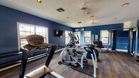 Located at 4390 Clearwater Way in Lexington, Kentucky, this single-floor fitness center is a well-equipped exercise facility designed to support a comprehensive workout experience. The space is immediately defined by its bold, deep blue walls contrasted against crisp white trim and a bright white ceiling, creating an energetic and focused atmosphere. Multi-toned striped carpeting covers the floor throughout, adding texture and durability underfoot. The facility is outfitted with a range of cardio equipment, including treadmills, elliptical machines, and a stationary cycling bike, offering users varied aerobic training options. A dedicated strength training area features a cable machine, adjustable bench, and a dumbbell rack, providing ample resources for resistance workouts. A full-length wall mirror with a wooden ballet-style barre runs along one wall, expanding the perceived depth of the room while also accommodating stretching and functional fitness routines. Multiple windows are positioned throughout the space, inviting abundant natural light and offering views of the surrounding outdoor landscape. Wall-mounted flat-screen televisions are positioned for visibility from key workout stations. Ceiling fans and recessed lighting ensure comfort and visibility during any time of day, making this fitness center a practical and well-appointed amenity.