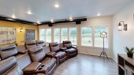 This single-floor screening and entertainment room is thoughtfully designed to deliver an immersive media experience with a comfortable, cinematic atmosphere. The centerpiece of the space is a curved arrangement of plush brown leather recliner seats, complete with built-in cup holders, providing generous seating capacity for groups. A wall-mounted flat-screen television sits atop a media console along one wall, flanked by wall sconces that cast warm, ambient light throughout the room.

The design blends industrial and modern touches seamlessly. Exposed black ductwork runs along the ceiling, adding a distinctive character, while recessed lighting ensures the space is well-illuminated. One accent wall is painted in a bold golden yellow, adorned with vintage-style theater posters and signage, reinforcing the room's cinematic theme. A retro-style floor spotlight lamp adds to the theatrical aesthetic.

A bank of large windows along one wall floods the space with natural light and offers pleasant views of the surrounding greenery and open landscape, creating an airy feel. Light wood-tone flooring completes the look, providing a clean contrast to the rich leather seating. A small café-style table and chairs occupy a corner, offering an additional gathering spot. A glass-paneled door provides easy access to the adjoining areas of the building.