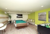 This well-appointed game room offers a versatile entertainment space designed for both active recreation and relaxed socializing. The room is anchored by a full-sized pool table with a teal felt surface, positioned centrally beneath pendant lighting and recessed ceiling fixtures that provide ample illumination throughout the space. Adjacent to the pool table, a foosball table occupies its own dedicated area, set against a yellow-green accent wall adorned with a pair of large, textured metallic artworks.

A sweeping landscape mural dominates one wall, depicting a serene, warm-toned horizon that adds depth and visual interest to the room. Beneath it, a pair of round white bistro tables with modern white chairs offer casual seating options. The light hardwood flooring runs throughout, contributing to a clean, contemporary aesthetic that ties the various zones together cohesively.

A dedicated lounge area features a sectional sofa with coordinating accent pillows, positioned facing a wall-mounted flat-screen television — ideal for gaming or media viewing. A sliding glass door provides access to an outdoor area, allowing natural light to filter into the space. The overall design balances energetic entertainment features with comfortable, inviting touches.