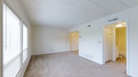 This single-floor space presents a straightforward and functional layout centered around a generously sized main room. The room is finished with wall-to-wall beige carpeting that adds a soft, neutral foundation throughout. White walls and a flat white ceiling contribute to a bright, clean aesthetic, while crown molding along the upper perimeter of the room provides a subtle architectural detail. A ceiling-mounted HVAC vent ensures climate control, and a baseboard heating unit is positioned along the lower wall, offering additional comfort options.

The room is open and unobstructed, providing flexibility for various furniture arrangements. An open doorway on the far wall leads to an adjoining space, suggesting additional rooms beyond the main area. To the right, a louvered closet door indicates built-in storage, adding practicality to the layout. Light switch plates are visible along the walls, and electrical outlets are positioned at standard intervals throughout.

Overall, the space maintains a simple, neutral palette with minimal ornamentation, creating a versatile environment that can easily be adapted to a variety of uses or personal styles.