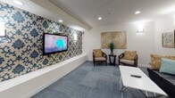 This thoughtfully designed single-floor amenity space offers a variety of functional areas, each styled with care and attention to detail. The open layout, visible from the dollhouse view, seamlessly connects distinct zones while maintaining a cohesive and inviting aesthetic.

The lounge area serves as a central gathering space, featuring a plush black leather sofa accented with teal and gold throw pillows, paired with carved wood accent chairs and a sleek white coffee table. Framed artwork and wall sconces lend a polished, curated feel, while blue-grey modular carpeting grounds the seating arrangement. A wall-mounted flat-screen television is set against a bold blue damask-patterned accent wall, creating a striking focal point.

Adjacent to the lounge is an intimate dining nook, distinguished by matching damask wallpaper and a substantial round wood dining table paired with contemporary chairs. A drum pendant light overhead adds warmth to the recessed-lit space.

The main open area features warm-toned hardwood flooring and includes a foosball table, offering a recreational element ideal for social gatherings. A wet bar or kitchenette with dark wood cabinetry and a stone countertop is positioned nearby, providing convenient amenities.

A glass-door fitness area is also accessible from the main space, housing exercise equipment. Throughout, recessed lighting, framed artwork, and a consistent warm-neutral palette create a welcoming and versatile environment.