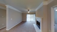This single-floor residence centers around a spacious living area designed for both comfort and functionality. The room is finished with crisp white crown molding that lines the perimeter, adding a refined architectural detail to the neutral beige walls. Plush wall-to-wall carpeting in a warm taupe tone covers the floor, contributing to the room's inviting atmosphere, while white baseboards provide a clean contrast.

A standout feature of the living area is the fireplace, framed by a classic white mantel with detailed millwork and surrounded by white ceramic tile, offering both a functional and decorative focal point. Adjacent to the fireplace, a sliding glass door fitted with vertical blinds allows natural light to filter into the space and provides access to an outdoor area with visible greenery beyond.

The ceiling is fitted with a ceiling fan and integrated light fixture, ensuring air circulation and ambient lighting throughout the room. The overall design is cohesive and understated, combining practical features with traditional finishes to create a comfortable, well-appointed living environment.