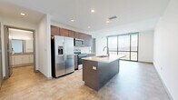 This thoughtfully designed one-bedroom, one-bathroom residence offers a refined modern aesthetic across a single floor. The open-concept kitchen serves as the heart of the unit, featuring rich dark-wood cabinetry, a white subway tile backsplash, stainless steel appliances, and a spacious center island with a built-in sink and light quartz countertop — ideal for both meal preparation and casual dining. Recessed lighting throughout keeps the space bright and inviting.

The living area flows naturally from the kitchen toward a dramatic floor-to-ceiling window wall framed in dark metal, flooding the interior with natural light and offering expansive elevated views of the surrounding cityscape. A structural concrete column adds an industrial architectural character to the open living zone.

The bedroom is generously proportioned and benefits from its own large window, continuing the theme of natural light and city views. A dedicated walk-in closet features built-in shelving and hanging rods, providing ample and organized storage.

The bathroom is clean and contemporary, with white subway tile lining a glass-enclosed walk-in shower, a floating vanity with neutral countertops, and a large mirror. Consistent tile flooring runs throughout the unit, unifying the space with a cohesive, polished look. A hallway connects the private spaces seamlessly to the main living area.
