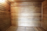 This single-level sauna is fully clad in horizontal dark wood planking, lending the interior a warm, enveloping atmosphere. The rich, deeply toned timber lines every wall surface, creating a cohesive and intimate environment conducive to relaxation. The wood has developed a characteristic patina, indicative of a well-used and carefully maintained space.

A solid timber door, fitted with a traditional wooden handle, provides entry to the sauna. A sliver of light along the door's edge hints at the contrast between the dimly lit interior and the outside environment, adding to the sense of seclusion within.

The flooring features slatted wooden grating, a functional and characteristic element of sauna design that promotes airflow and drainage while maintaining the all-wood aesthetic. A small stool or bucket is visible in one corner, consistent with traditional sauna use.

Subtle overhead lighting casts a soft, warm glow across the wooden surfaces, enhancing the dark tones of the timber and contributing to the calm, meditative ambiance. Overall, the space is defined by its consistent use of natural wood materials and its focus on creating a private, thermally efficient retreat.